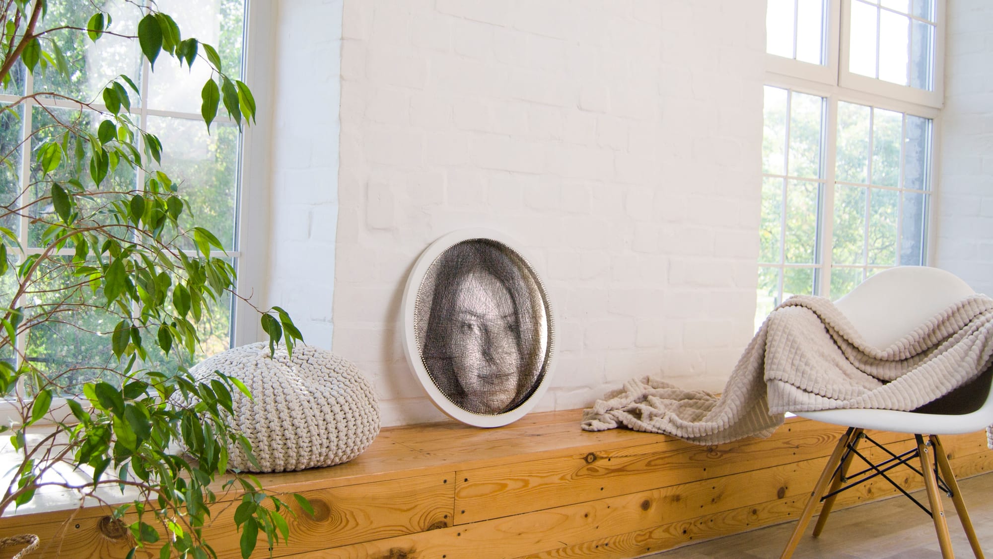 Round string art portrait leaning against a white brick wall on a wooden bench near a window and plants.