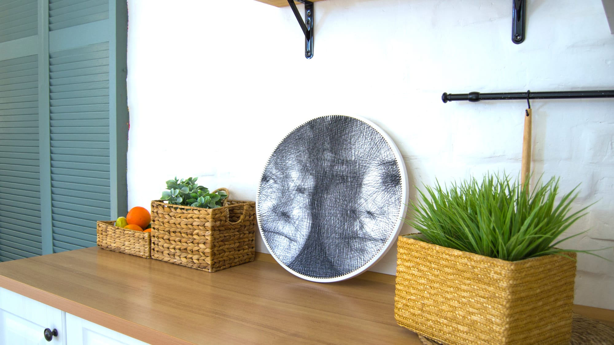 Circular string art portrait displayed on a wooden kitchen countertop between woven baskets and green plants.