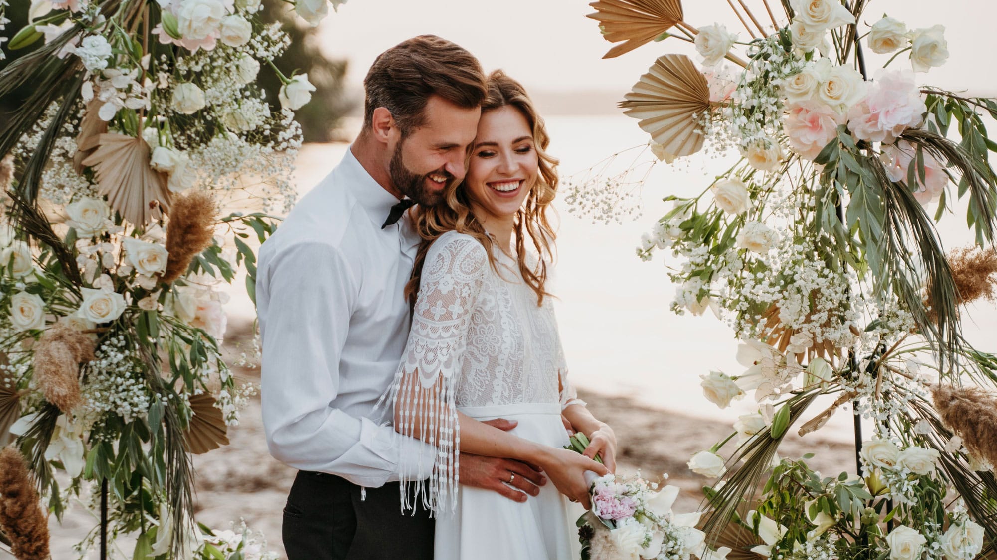 Smiling bride and groom standing under a decorative floral wedding arch near water, embracing and laughing during an outdoor ceremony.