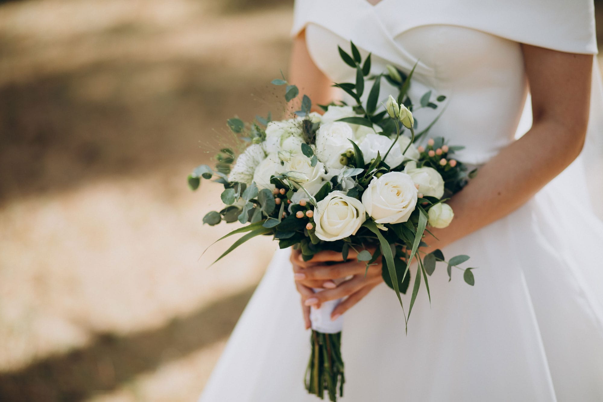 Close-up of a bride in a white wedding dress holding a bouquet of white roses and greenery outdoors.