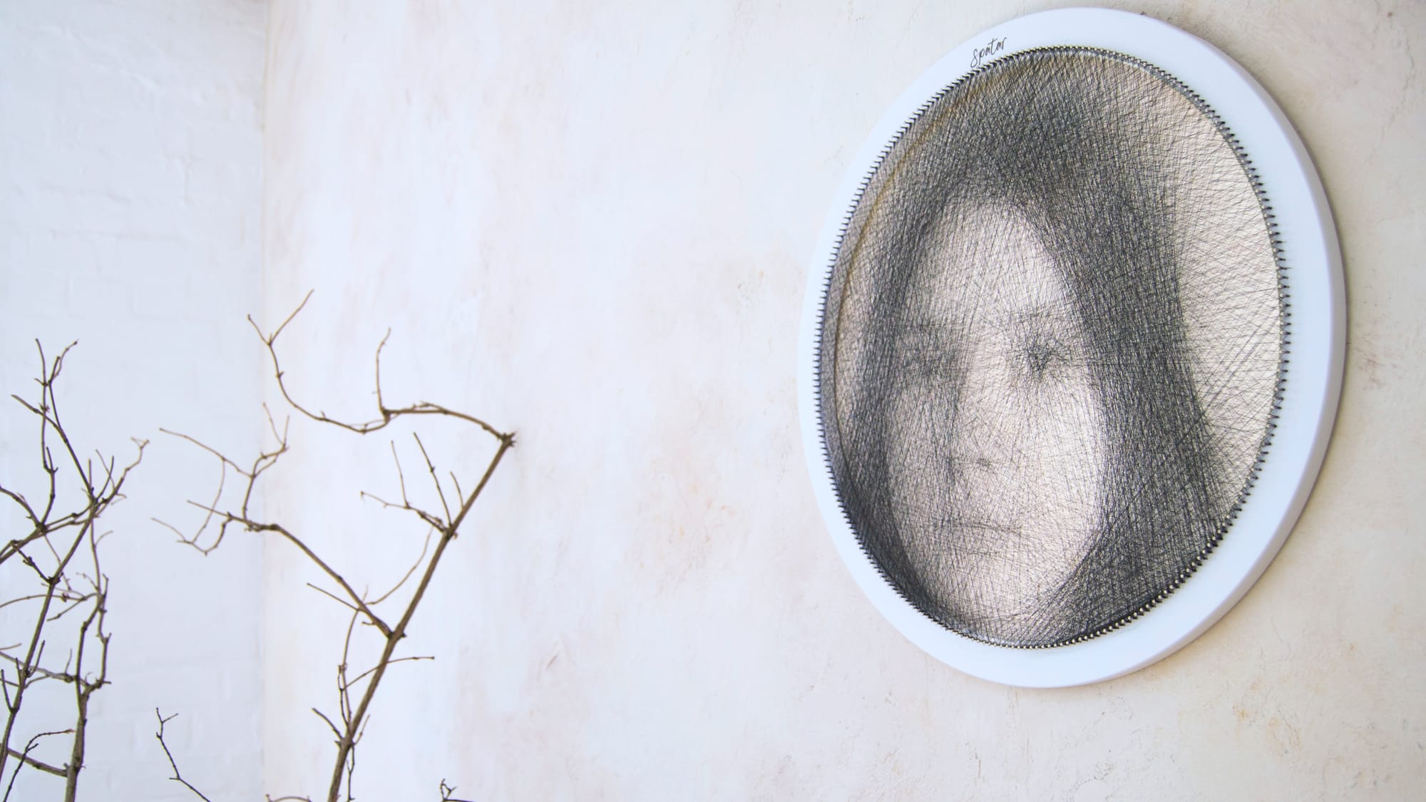 Round string art portrait made with fine black thread on a white circular frame, hanging on a light wall with a dried branch nearby.