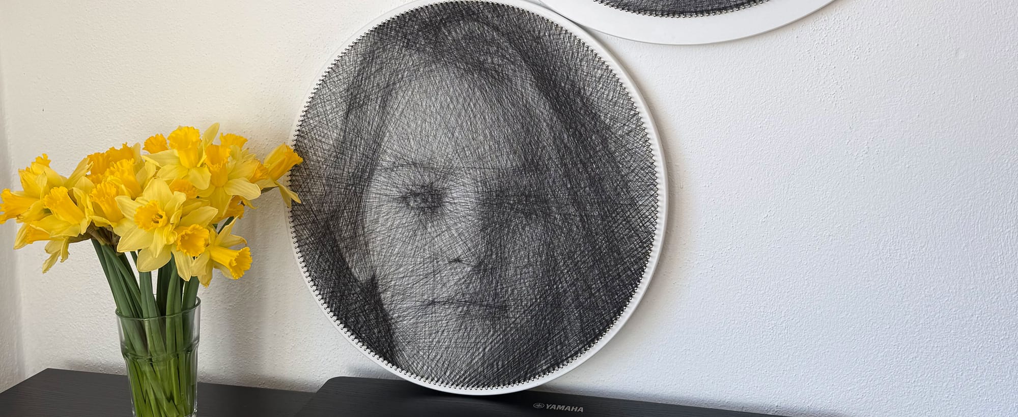 Close-up of a circular string art portrait made with black thread, displayed above a piano with flowers nearby.